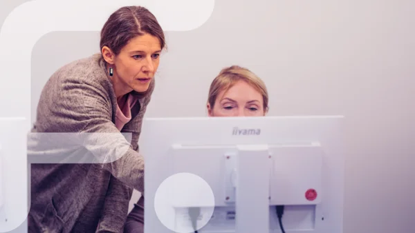 Zwei Frauen konzentrieren sich auf einen Computerbildschirm, eine steht und zeigt auf den Bildschirm, die andere sitzt davor. Im linken Bildbereich ist grafisch das stilisierte "F" des Projekts „Fempower“ eingeblendet.